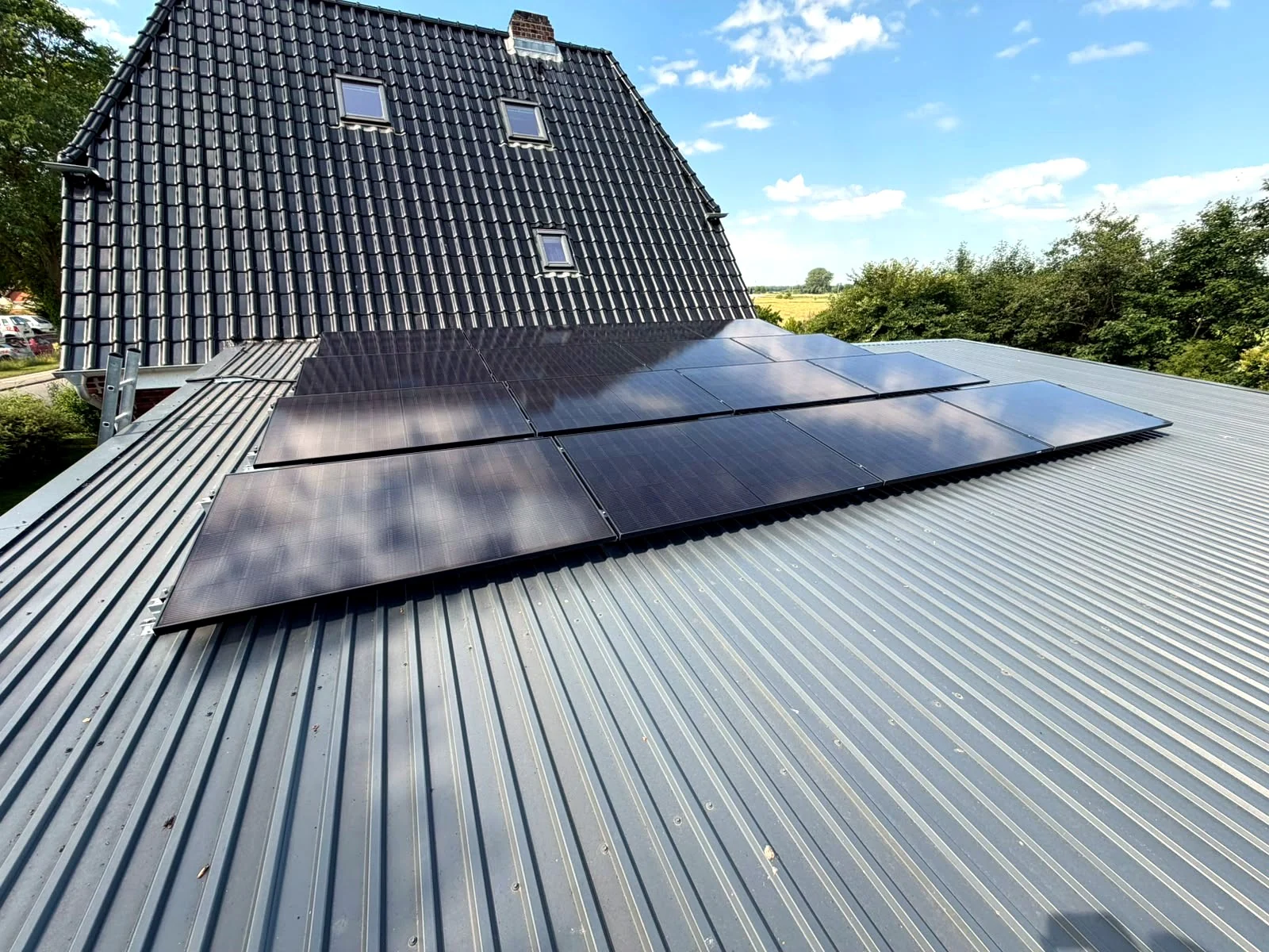 Photovoltaikmodule auf einem flach geneigten Trapezblechdach vor einem Haus mit steilem Ziegeldach